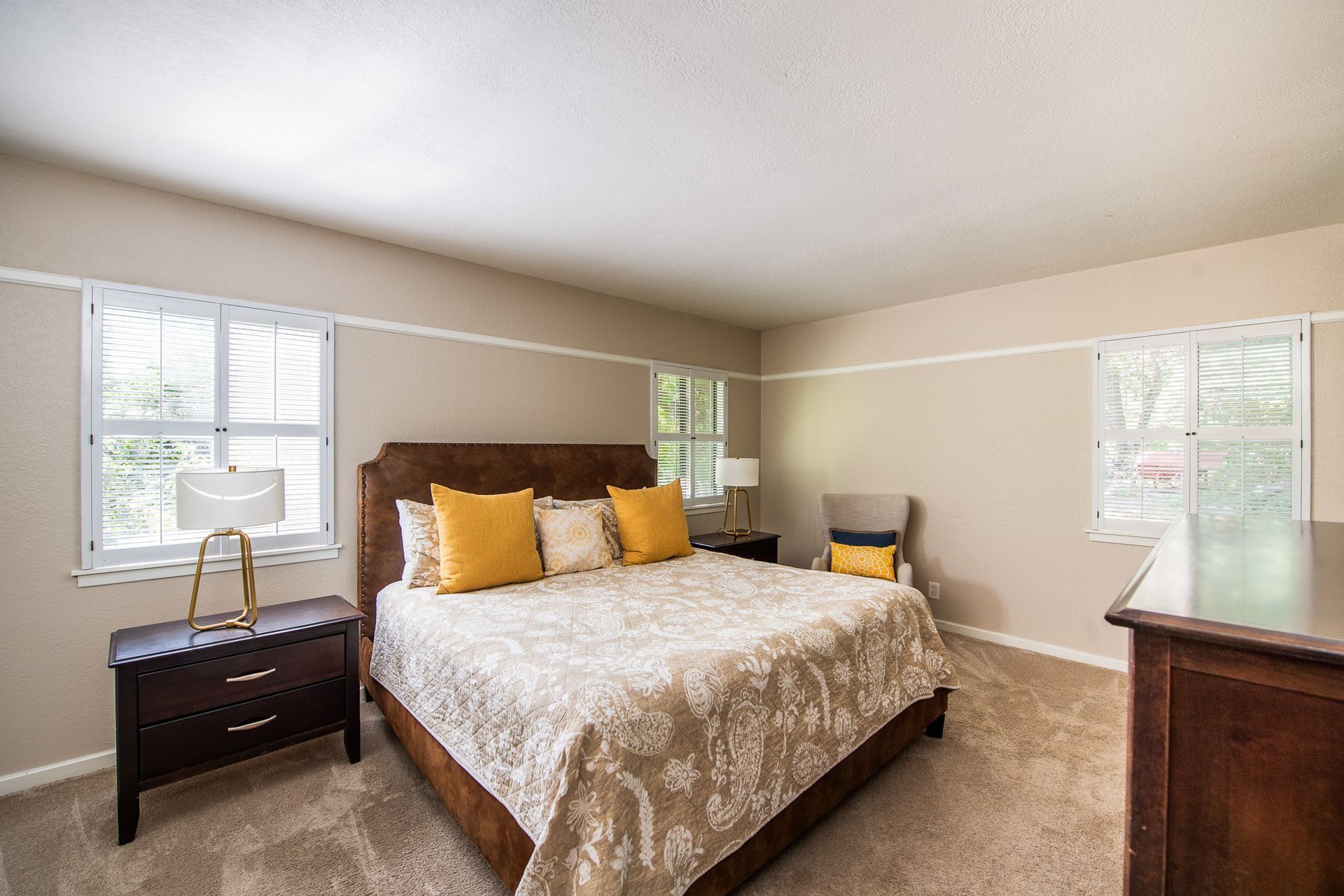 Spacious beige bedroom with a large bed, nightstands, windows, and carpeted floor