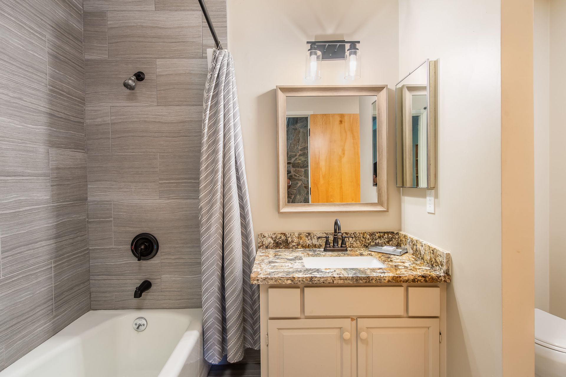 Bathroom with gray tiled shower, white tub, and granite vanity with mirror and light fixture