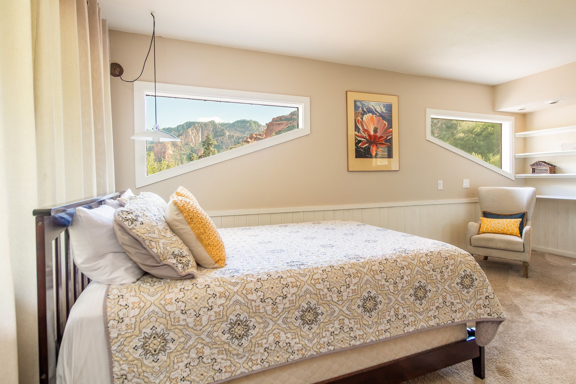 Bright bedroom with floral bedspread, two chairs, and large corner windows overlooking a hillside.