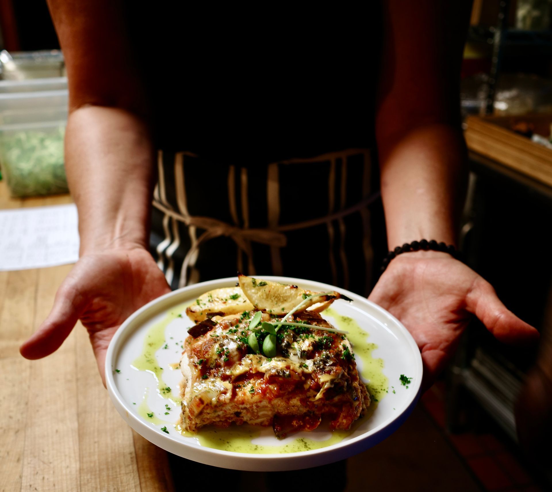 Hands presenting a plated savory dish topped with herbs and sauce on a wooden table