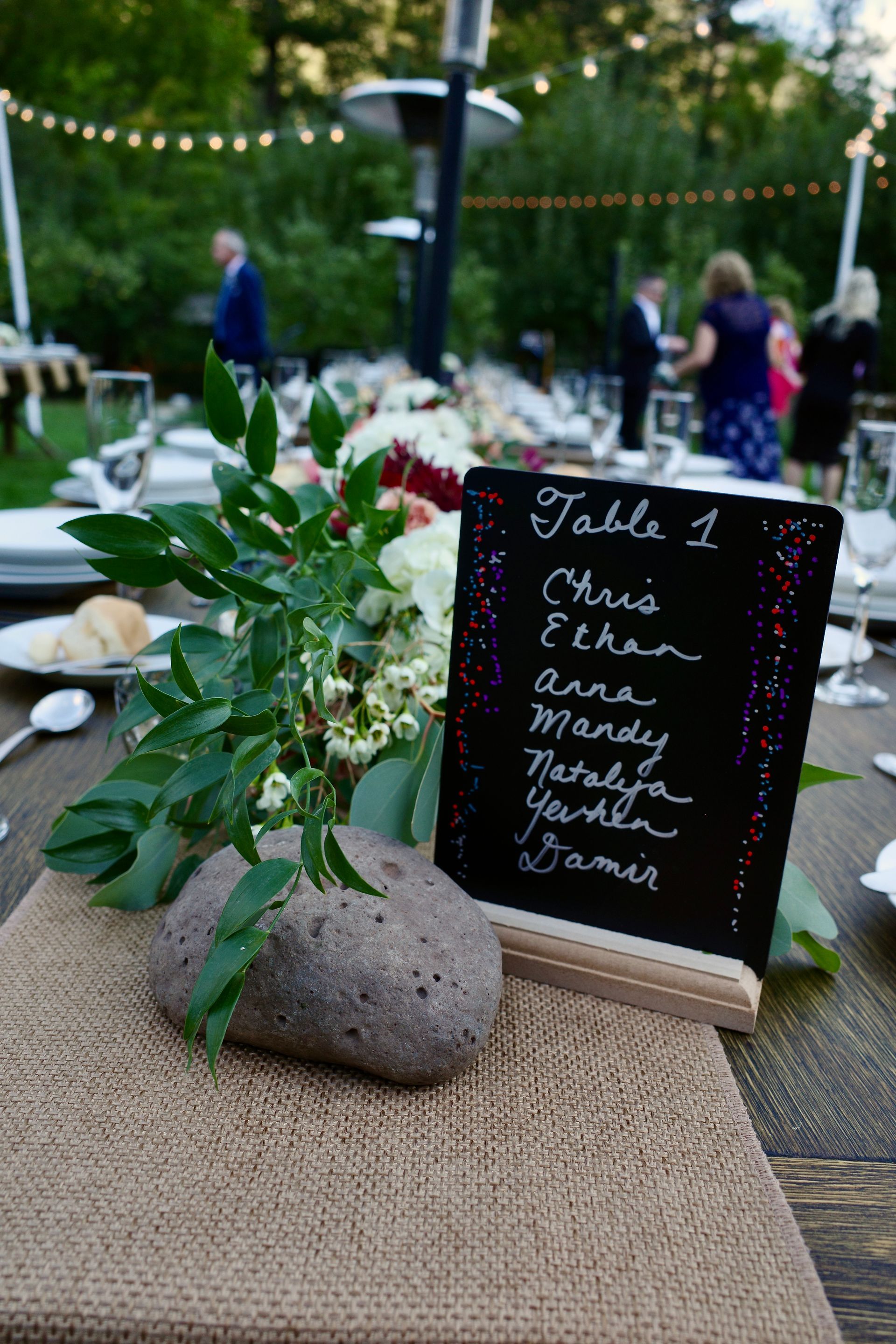 Table centerpiece with flowers, a chalkboard menu, and a stone weight on a burlap runner outdoors