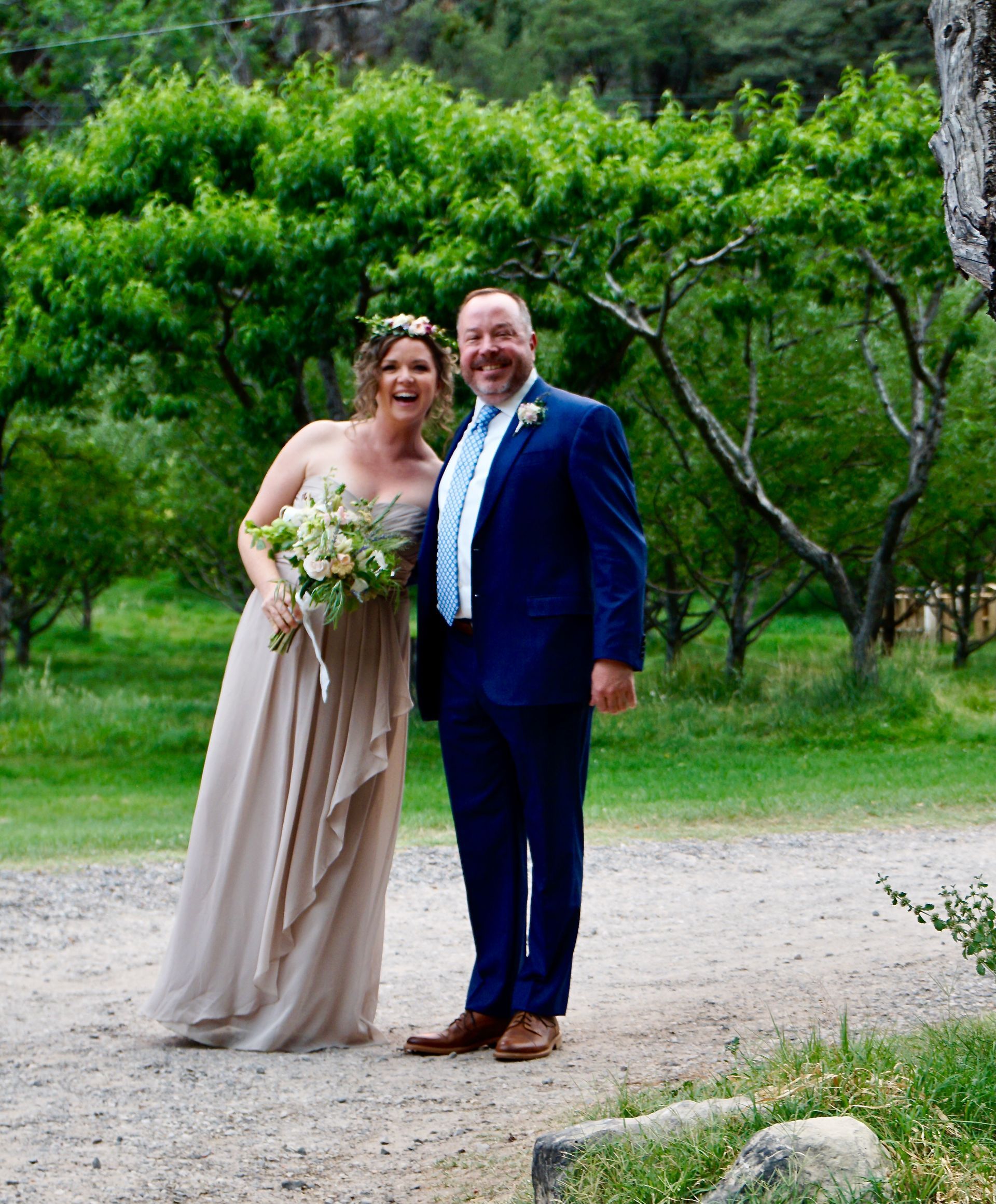 Smiling couple in wedding attire standing on a garden path with trees behind them