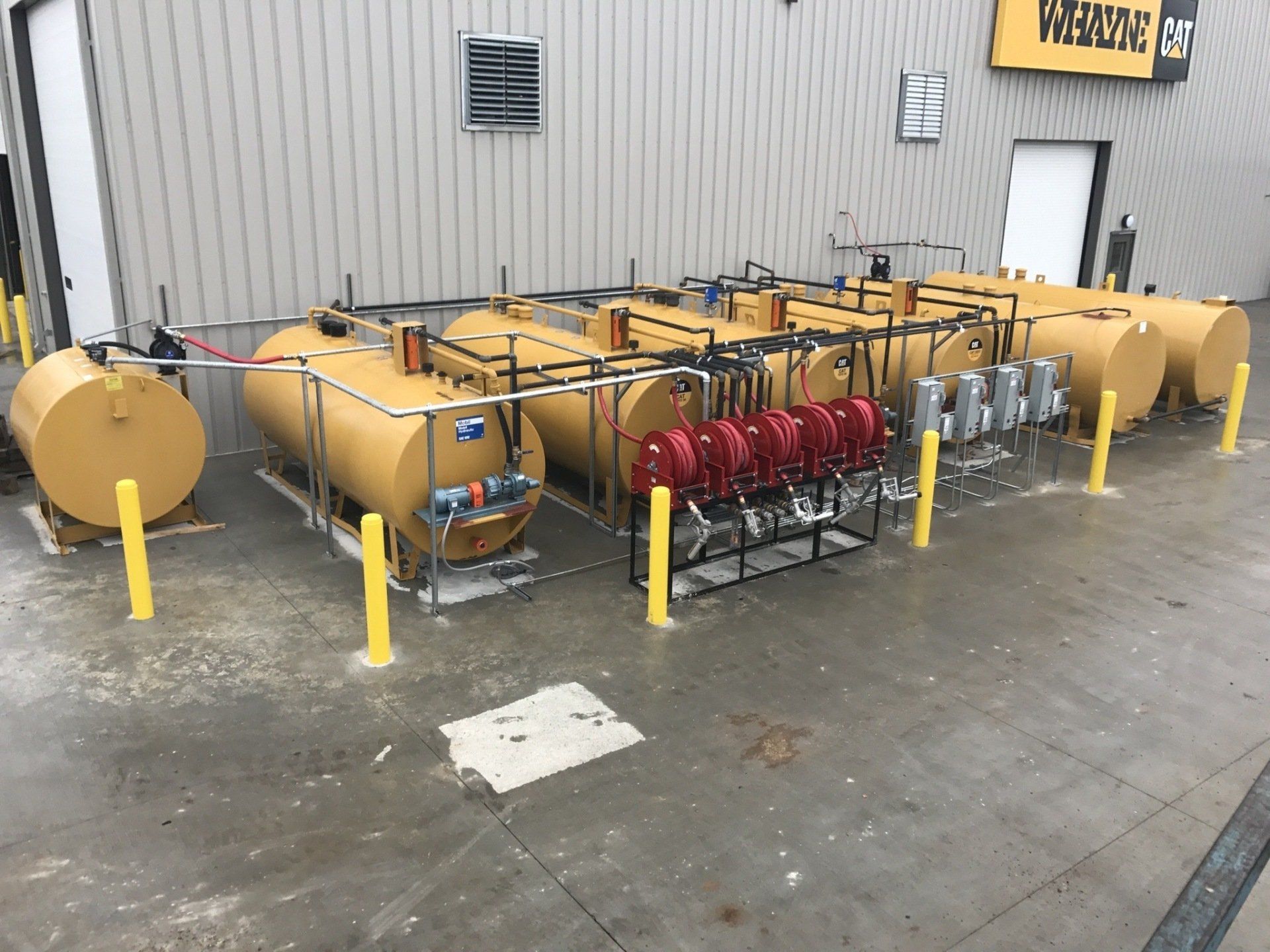 A row of yellow industrial fuel tanks with red hose reels outside a warehouse building.