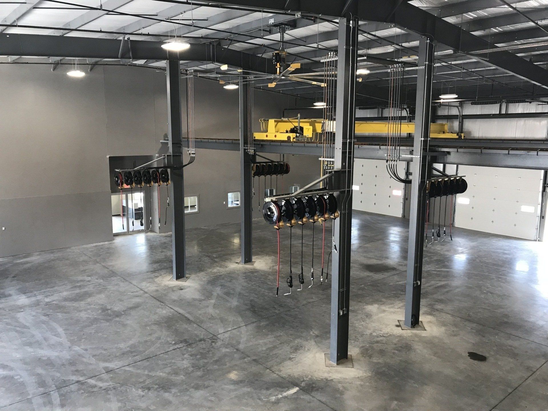 A wide, empty industrial warehouse with gray concrete floors, overhead service reels, and a yellow crane.
