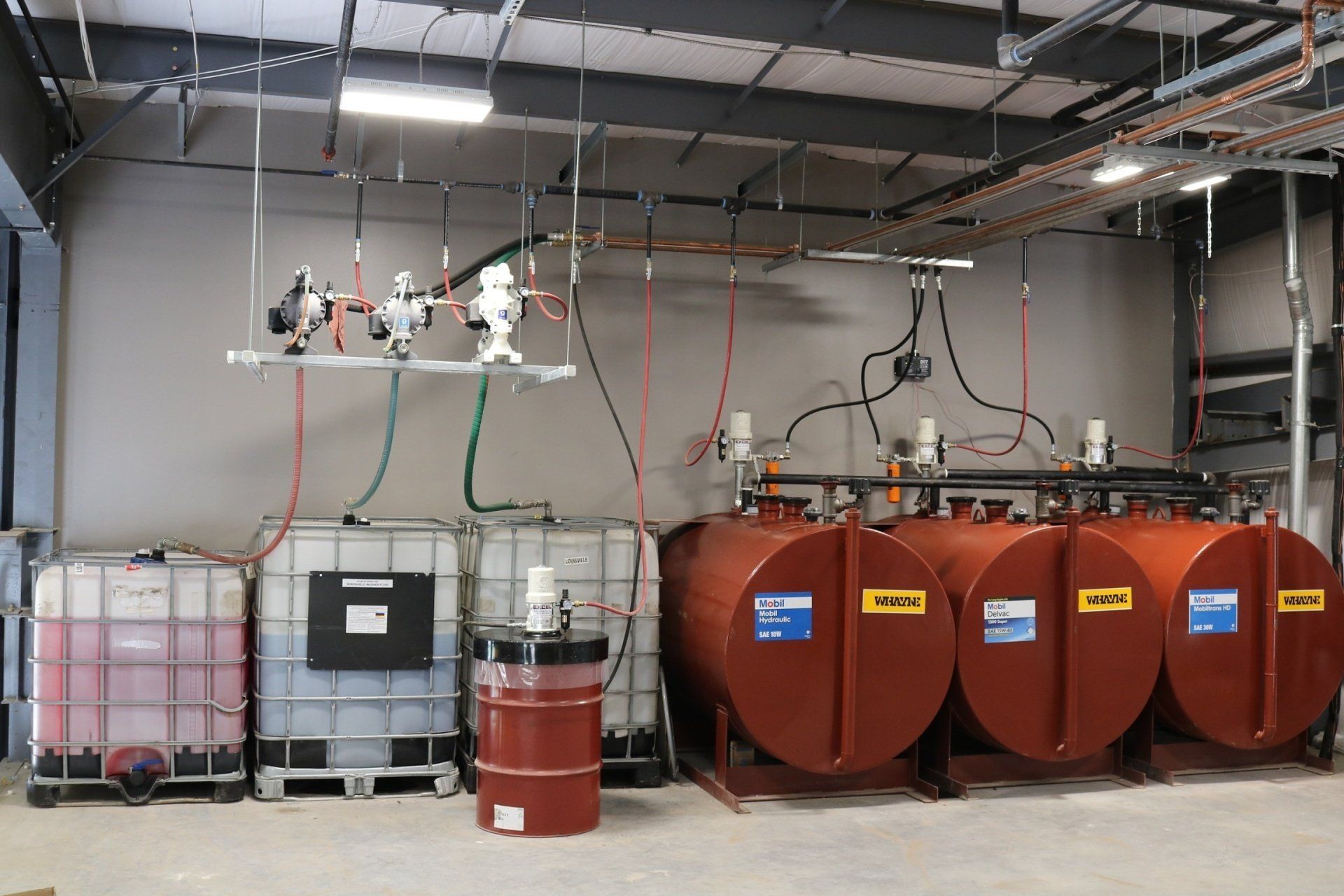 Industrial room featuring large liquid storage tanks, IBC containers, and a red barrel connected by overhead piping.