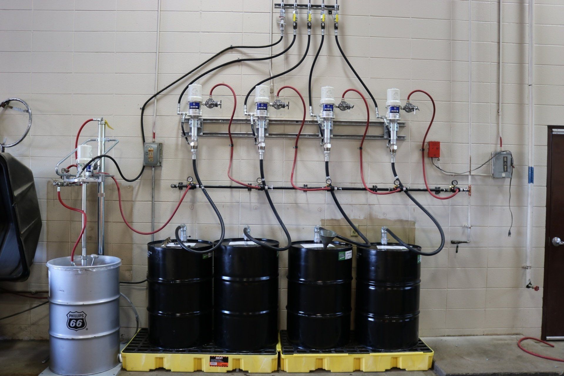 A wall-mounted pump system with four black barrels and one silver drum on yellow containment pallets in a workshop.