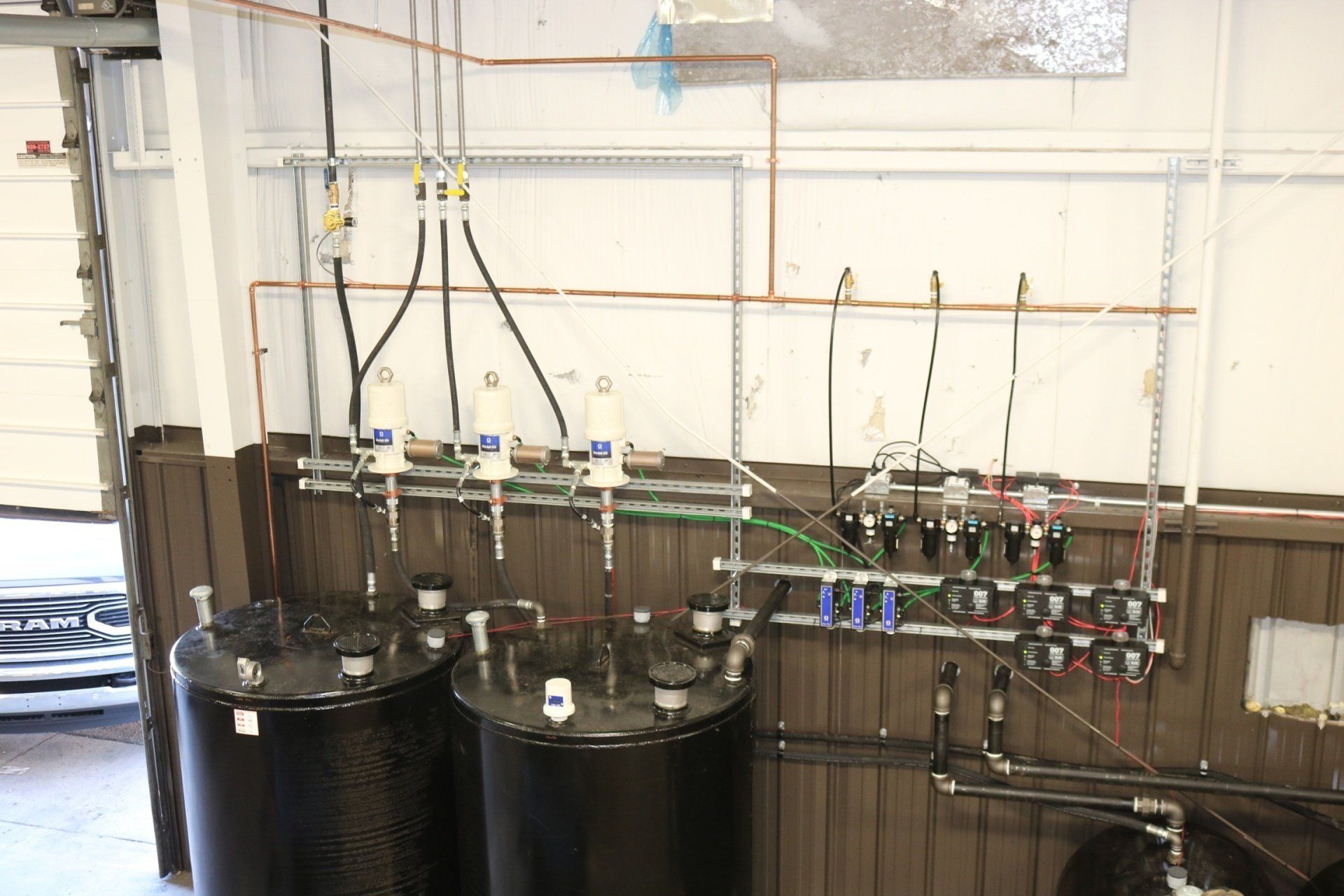 Two large black oil tanks connected to wall-mounted pumps, pipes, and control gauges in an industrial garage setting.