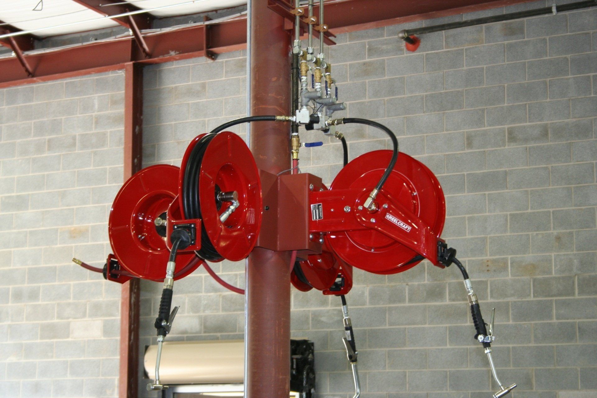 Red industrial hose reels mounted to a metal support column in a workshop with concrete block walls.