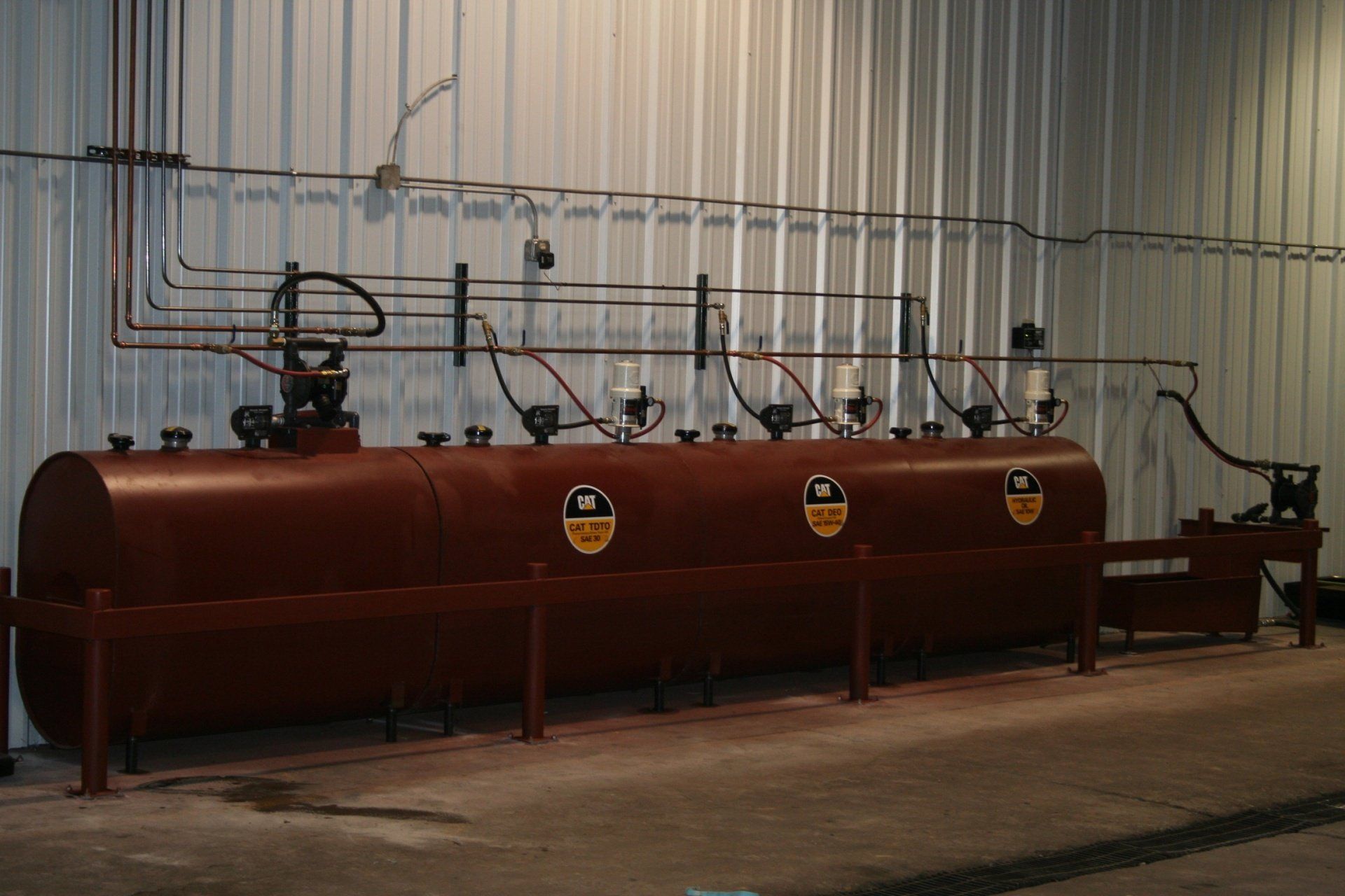 A horizontal brown industrial storage tank with multiple pipes and valves against a corrugated metal wall.