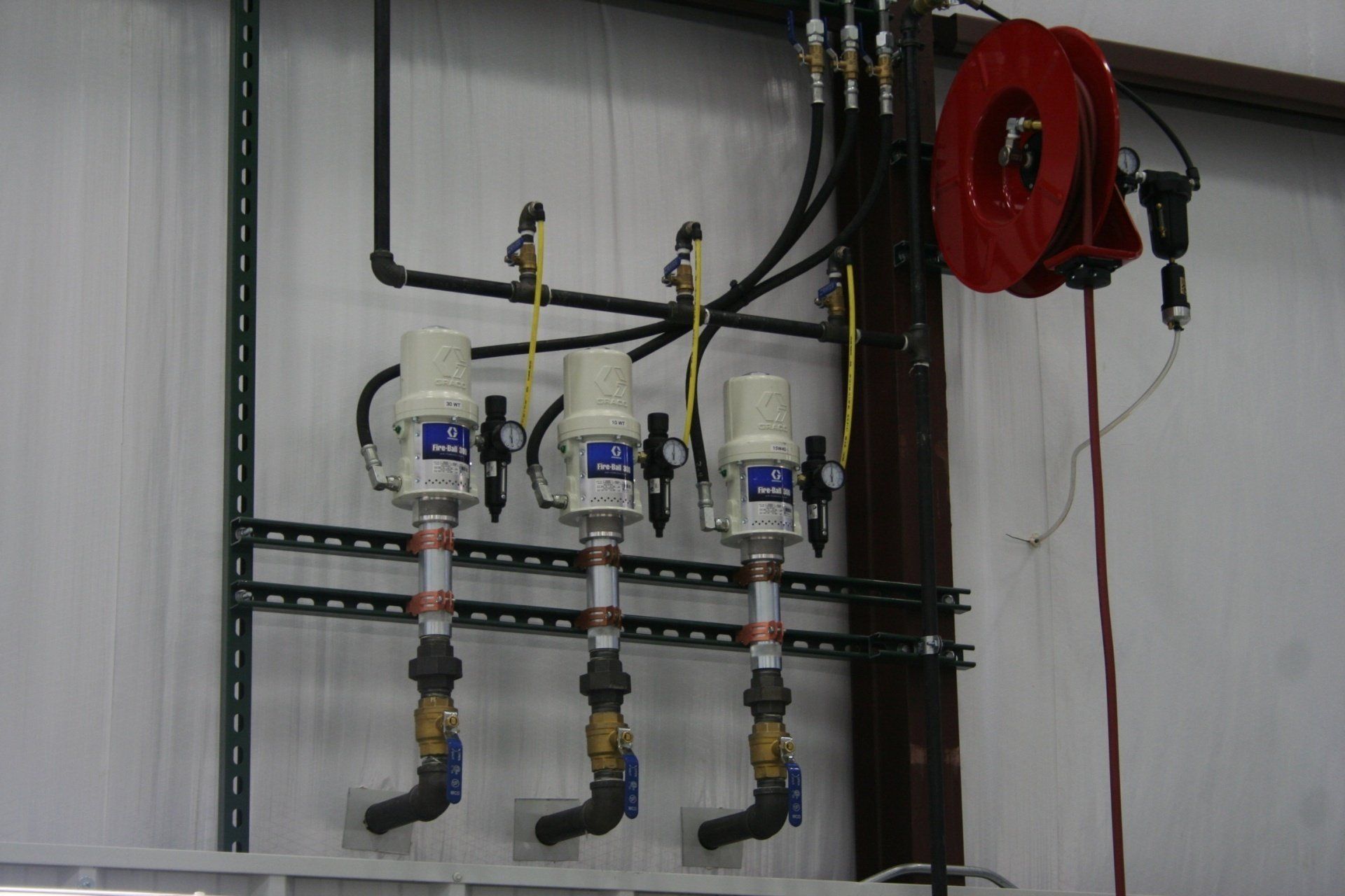 Three industrial fluid pumps mounted on a wall with plumbing, yellow hoses, and a red retractable hose reel.