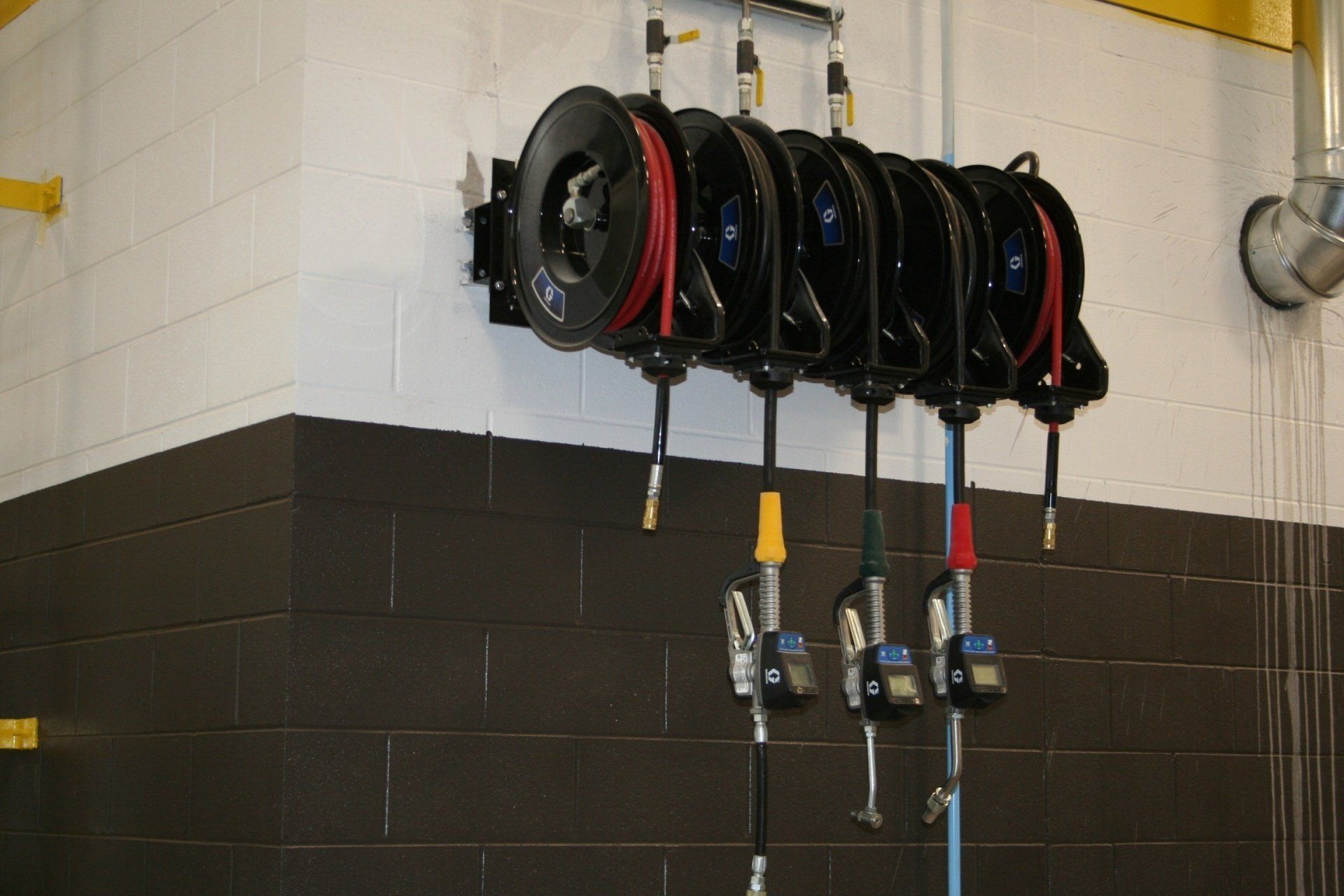Wall-mounted industrial fluid hose reels with dispensing nozzles in a workshop with white and dark gray brick walls.