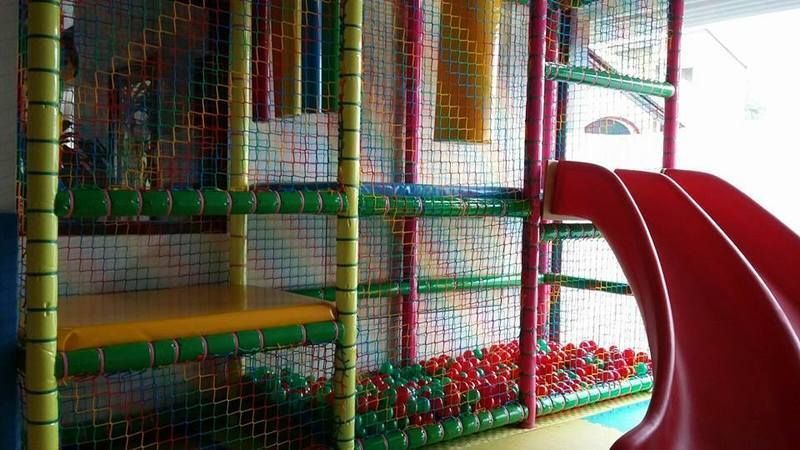 Area giochi interna per bambini con struttura di arrampicata a rete, piattaforme imbottite e uno scivolo rosso.