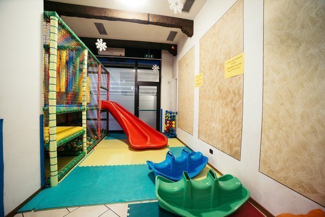 Sala giochi interna con scivolo rosso, giochi di arrampicata blu e verdi e tappeti imbottiti.
