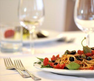 Piatto di pasta servito su un tavolo bianco con bicchieri da vino e posate in una sala da pranzo formale.