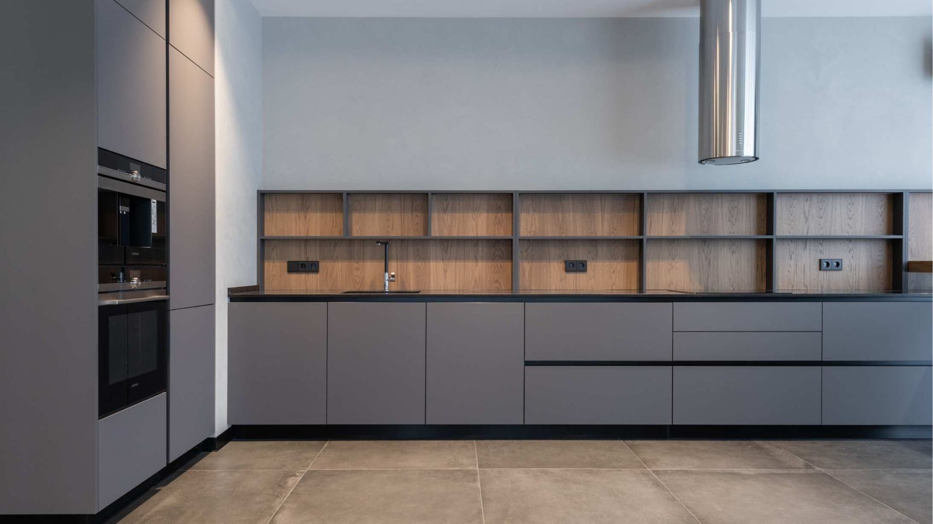 Modern minimalist kitchen with gray cabinets, wood shelves, built-in oven, and large tiled floor