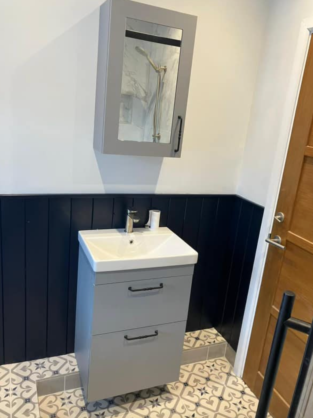 Small bathroom vanity with gray cabinet, white sink, mirror, navy wainscoting, and patterned floor tiles.