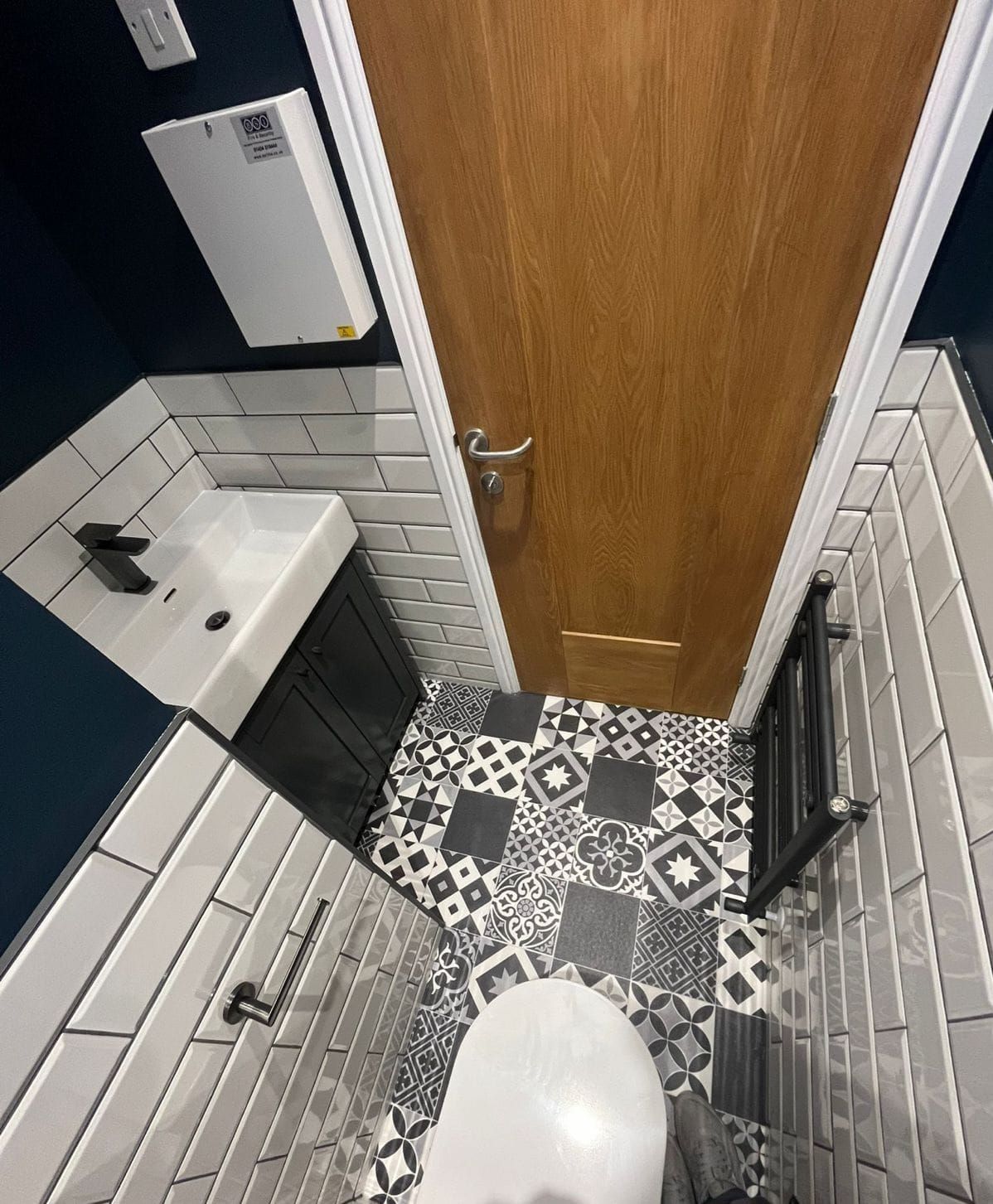 Narrow bathroom with toilet, sink, wooden door, and black-and-white patterned tile floor.