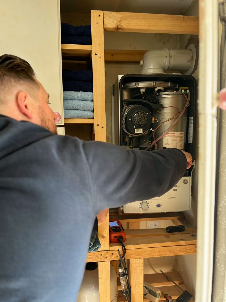 Person working on a white boiler with a screwdriver in a narrow closet next to shelves of folded linens