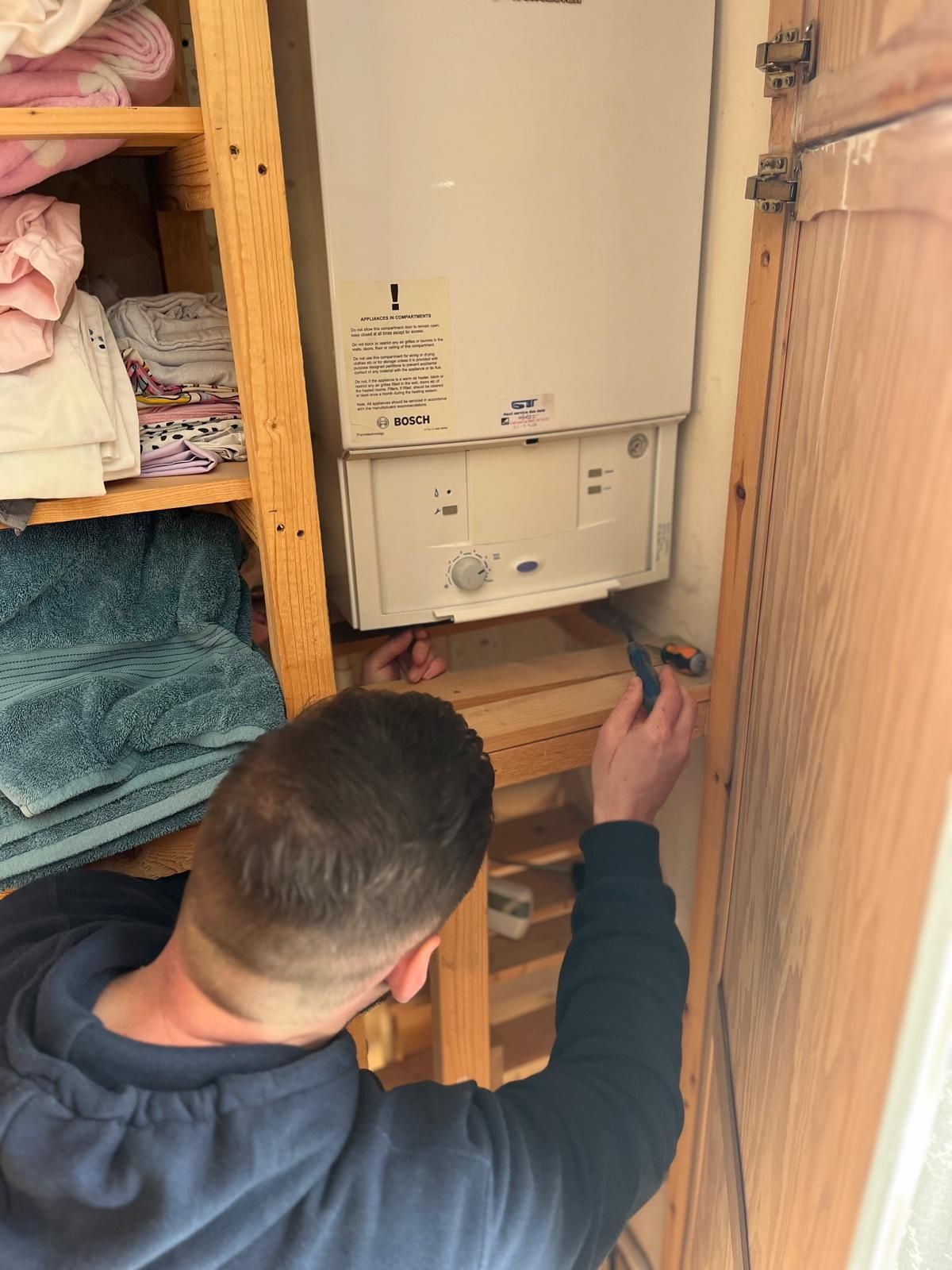 Person working on a white boiler with a screwdriver in a narrow closet next to shelves of folded linens