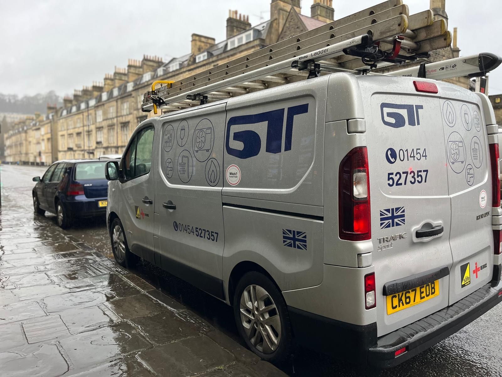 White GT van parked on a wet city street beside stone buildings, with another car ahead.