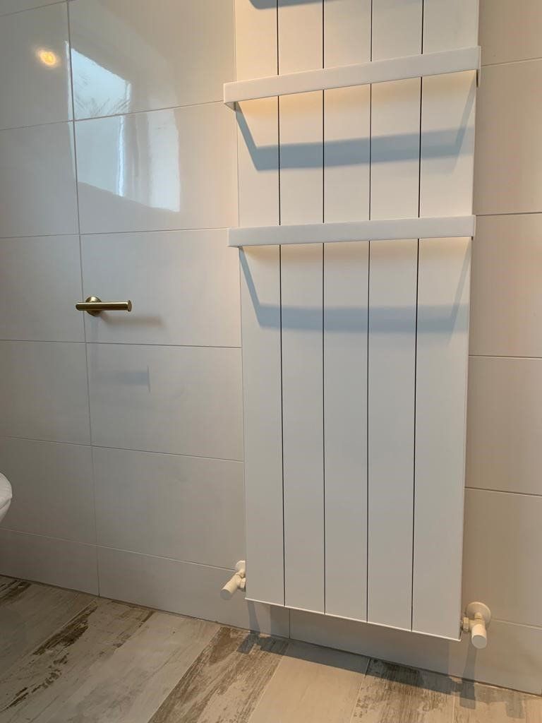 White bathroom towel radiator with shelves beside a tiled shower wall and gold door handle.