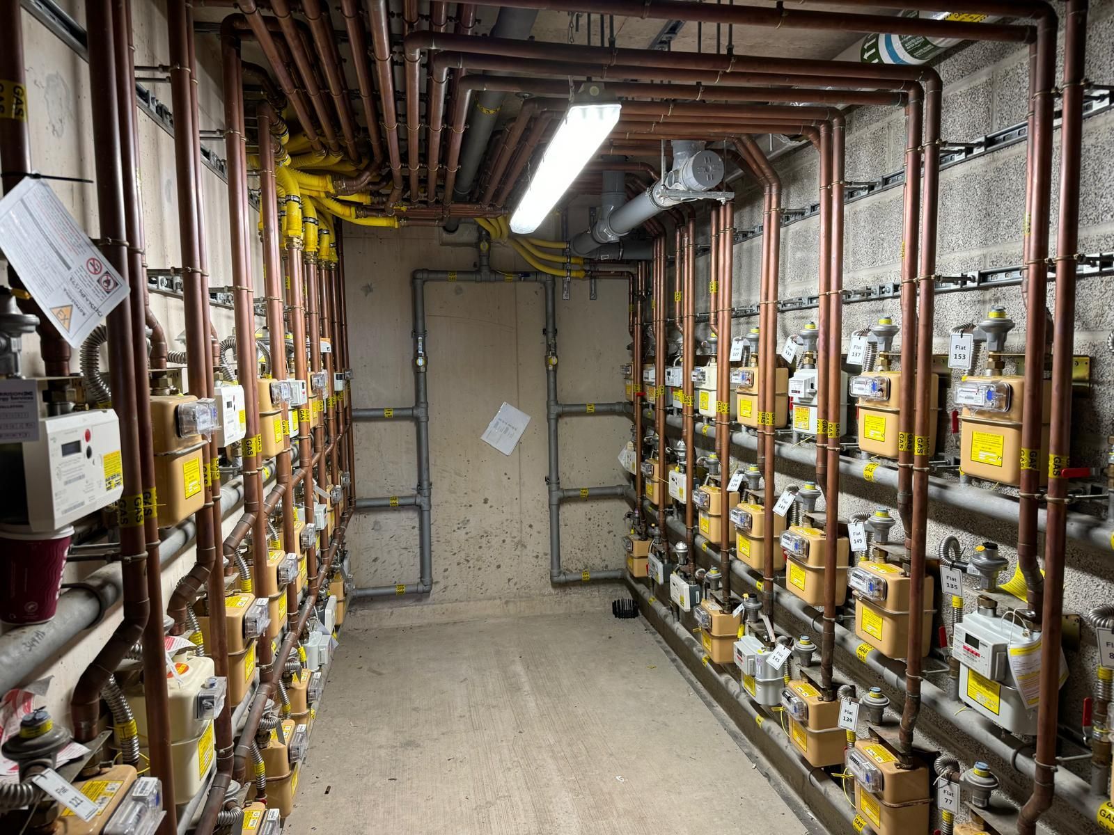 Narrow utility corridor with rows of gas meters and pipes along both walls, lit by fluorescent lights