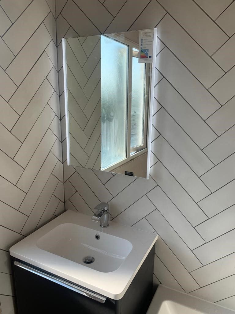 Small bathroom sink below a mirrored cabinet, with white tiled walls and a frosted window.