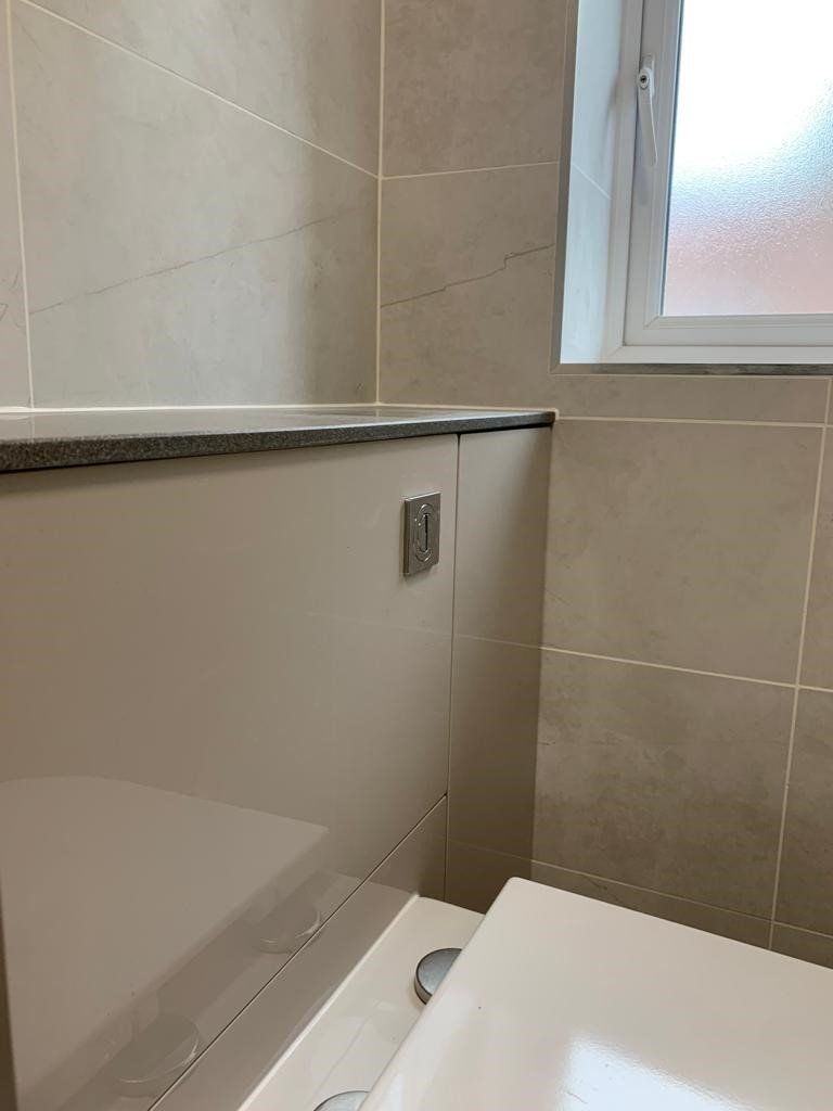 Bathroom corner with beige tiles, white toilet seat, and frosted window above a ledge.