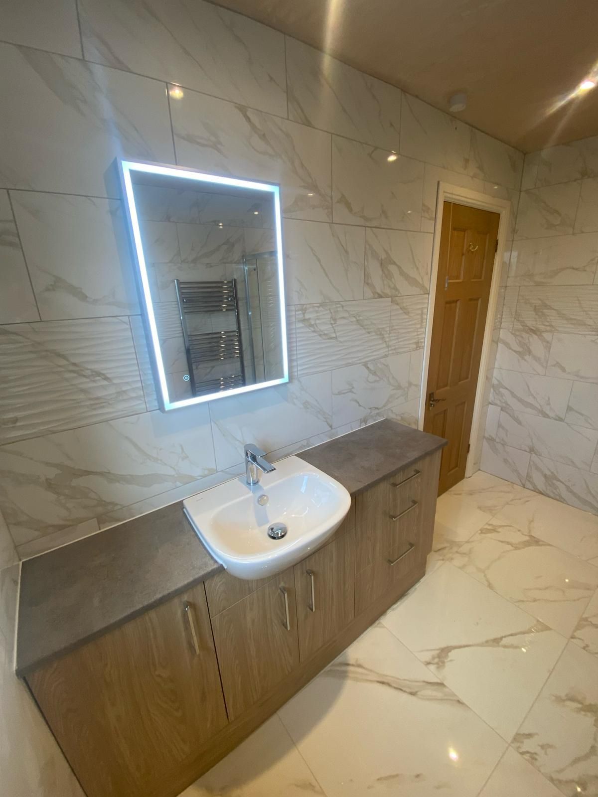 Modern bathroom vanity with lit mirror, white sink, wood cabinet, and marble-pattern tile walls and floor