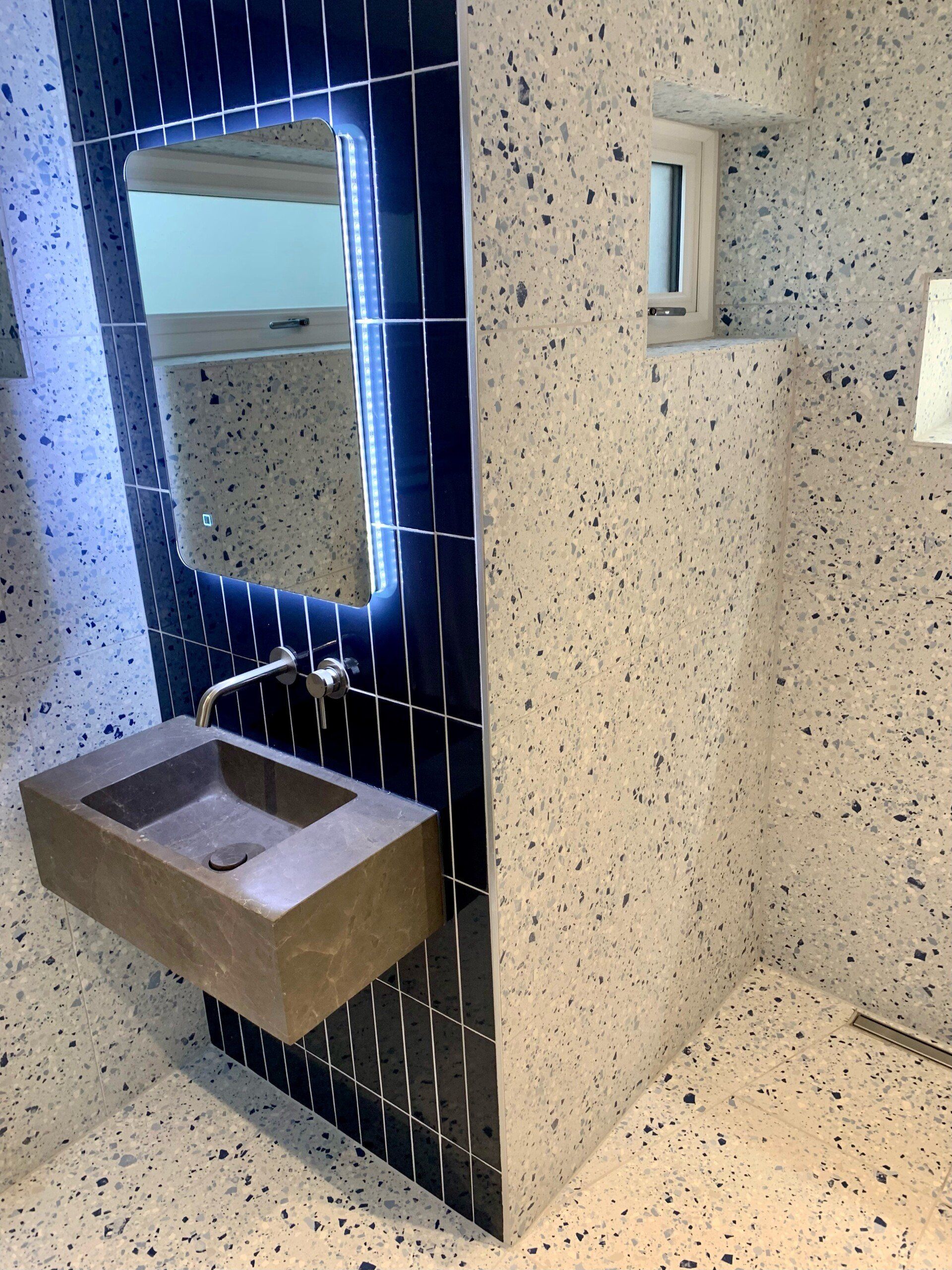 Small modern bathroom sink with mirror, blue tile accent wall, and speckled stone walls and counter