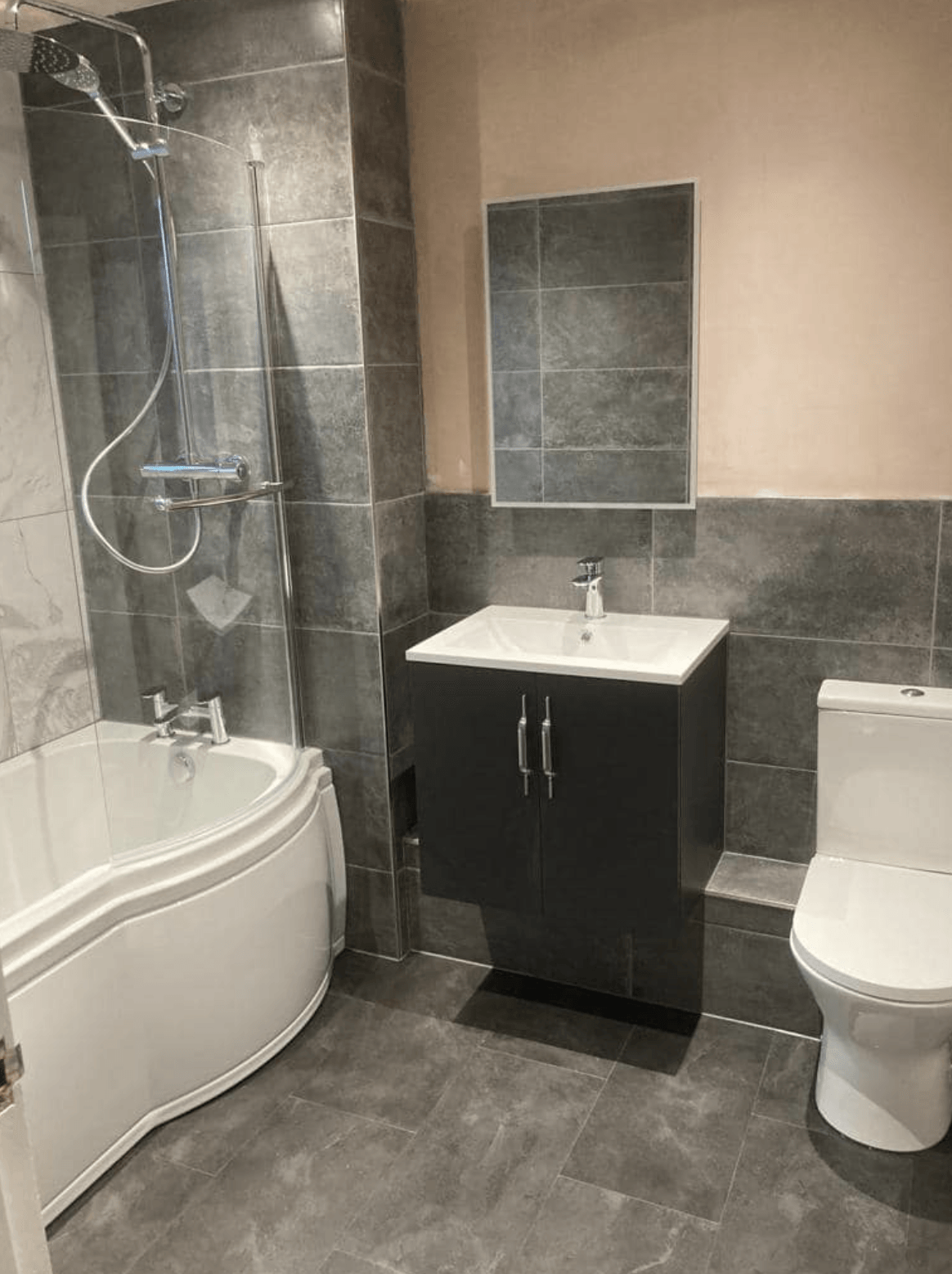 Small bathroom with bathtub shower, dark vanity, sink, mirror, and toilet on gray tile.