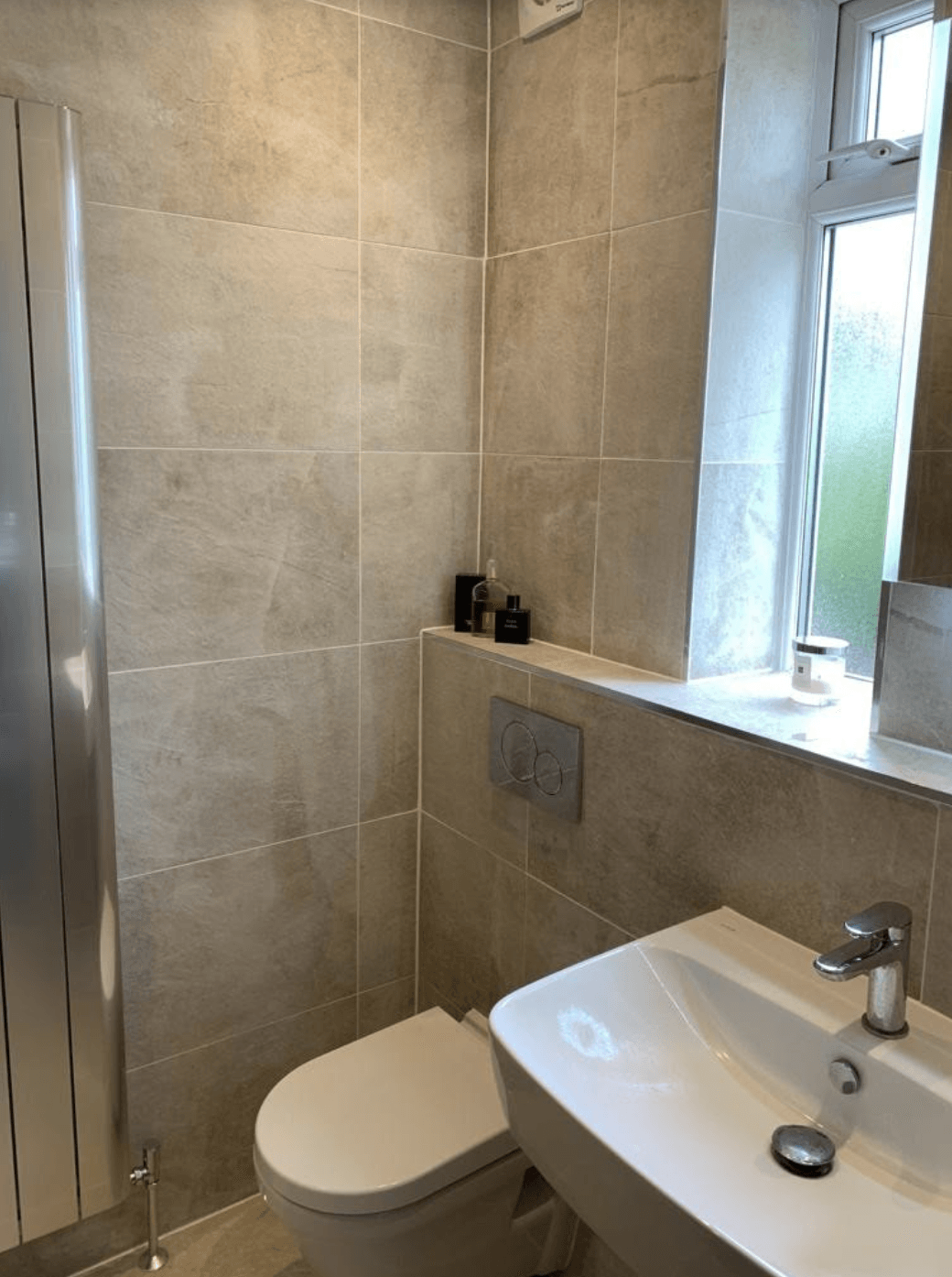 Small tiled bathroom with a toilet, sink, and window beside a glass shower door.