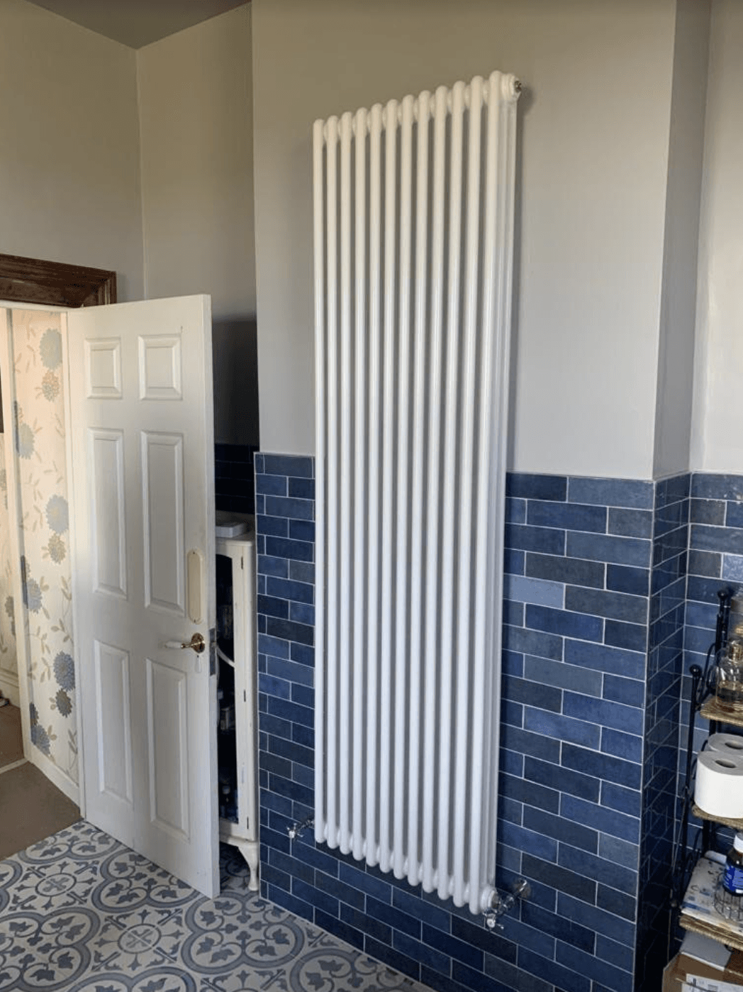 Tall white radiator on a gray wall above dark blue tiled wainscoting near an open door.