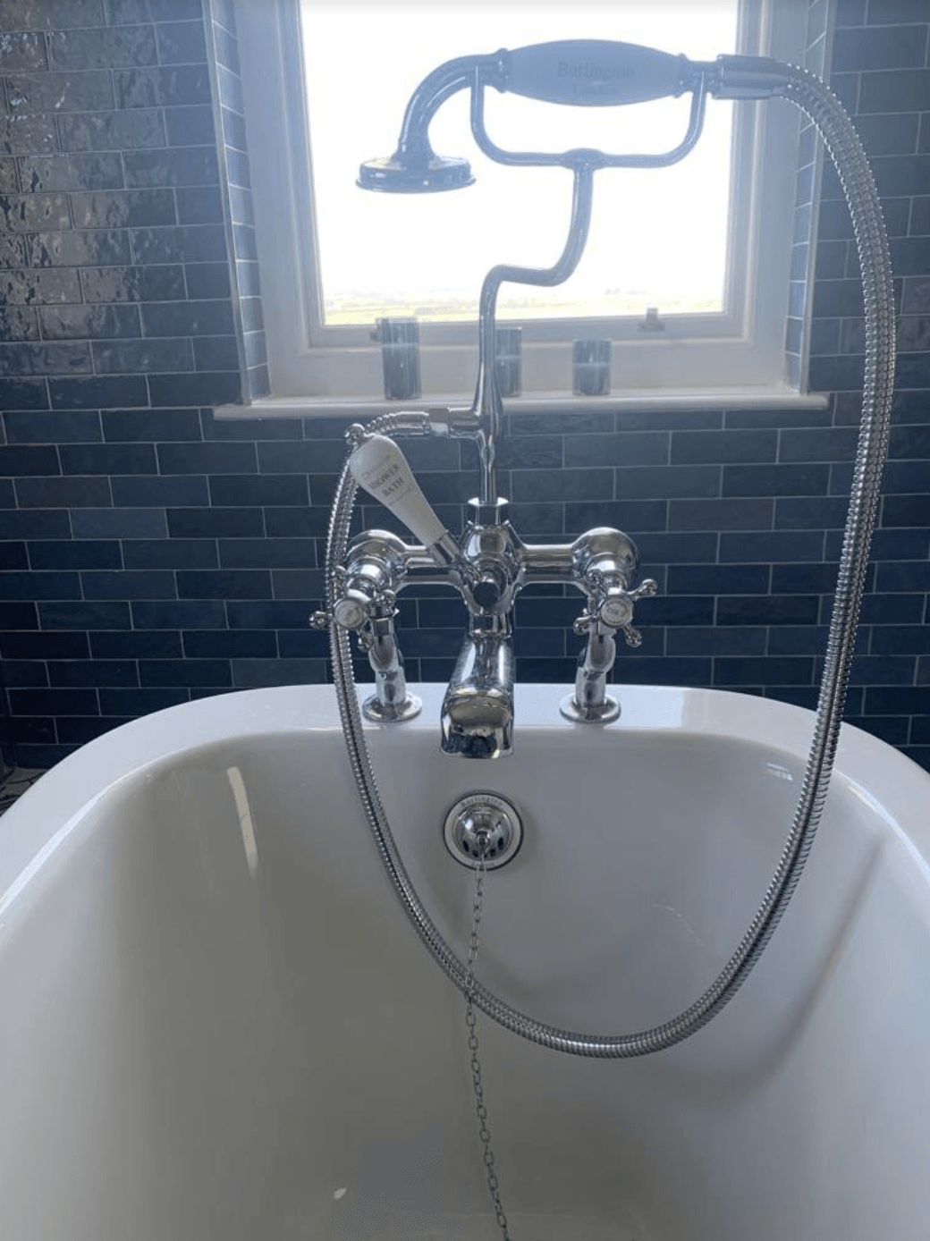 Freestanding bathtub with chrome faucet and handheld shower beneath a bright window and black tiled wall