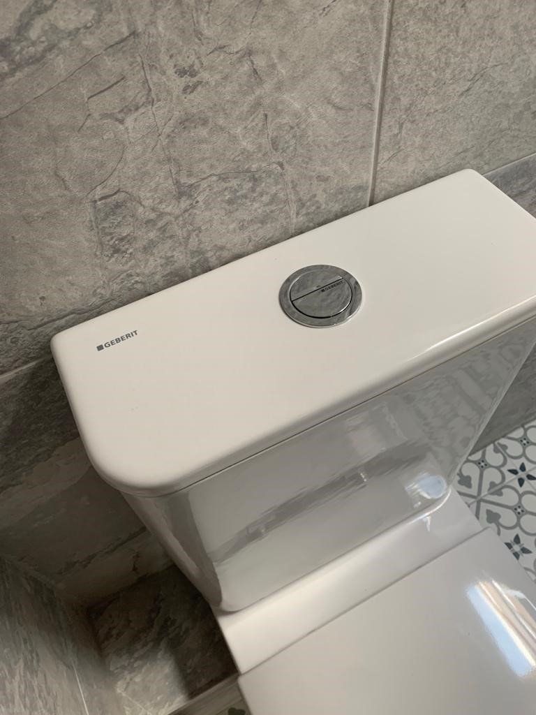 White toilet tank with a chrome flush button against a gray tiled wall and patterned floor.