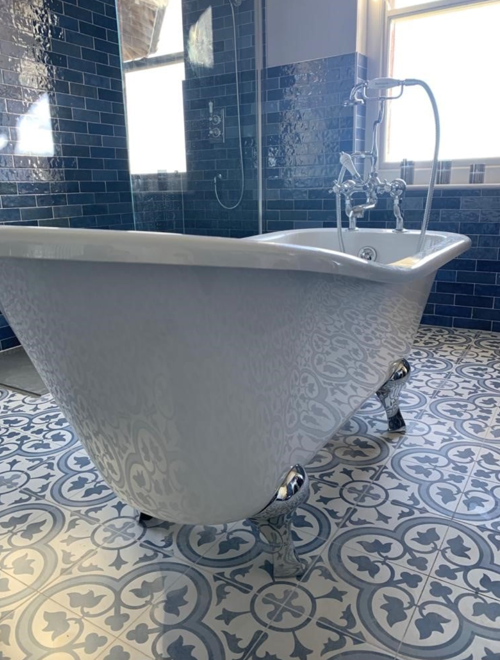 Freestanding white clawfoot bathtub in a blue-tiled bathroom with patterned floor tiles