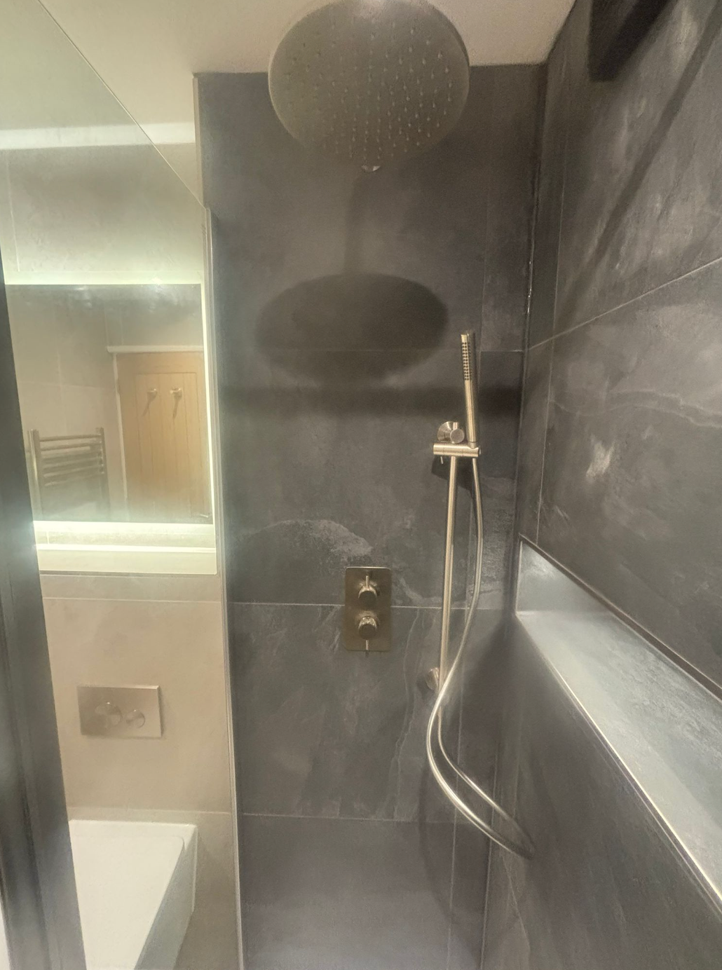 Modern bathroom shower with dark tile, rainfall head, handheld shower, and bathtub edge visible