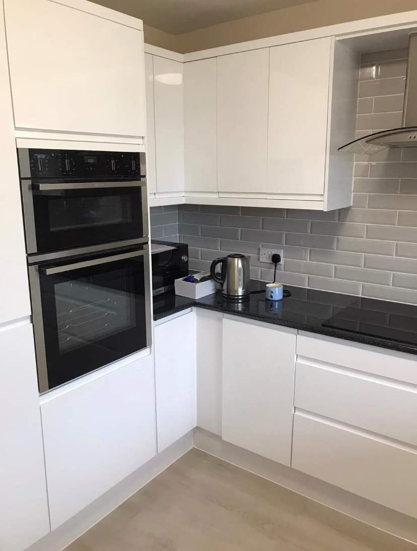 Modern white kitchen corner with built-in oven, black countertop, gray tiled backsplash, and stainless kettle.