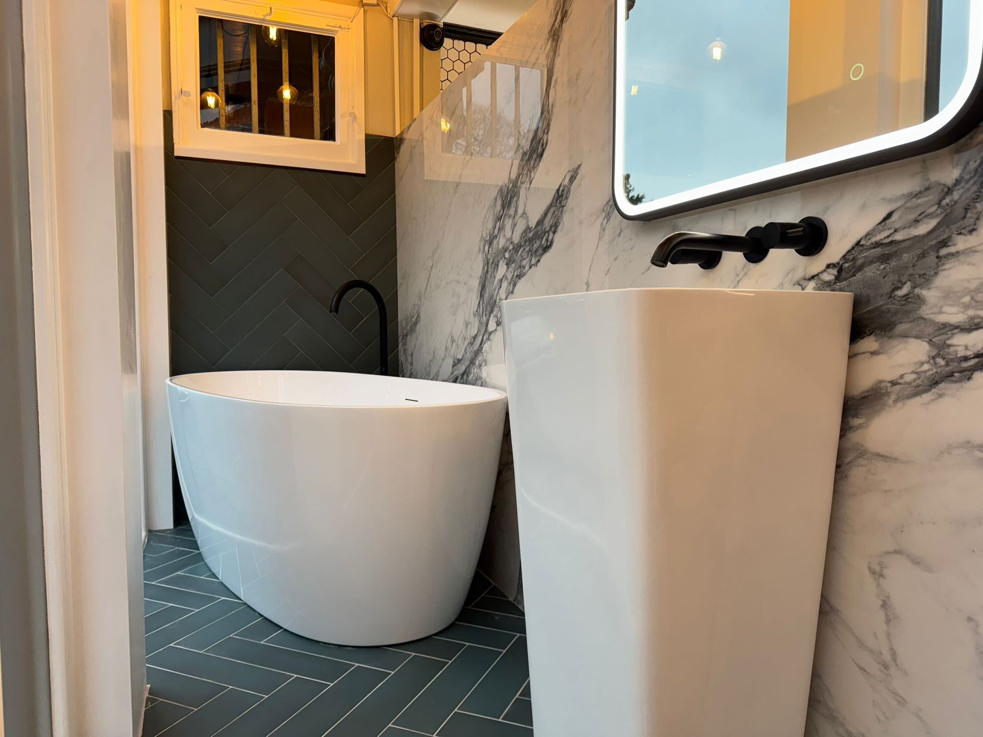 Modern bathroom with a freestanding tub, pedestal sink, marble walls, and a window above the tub