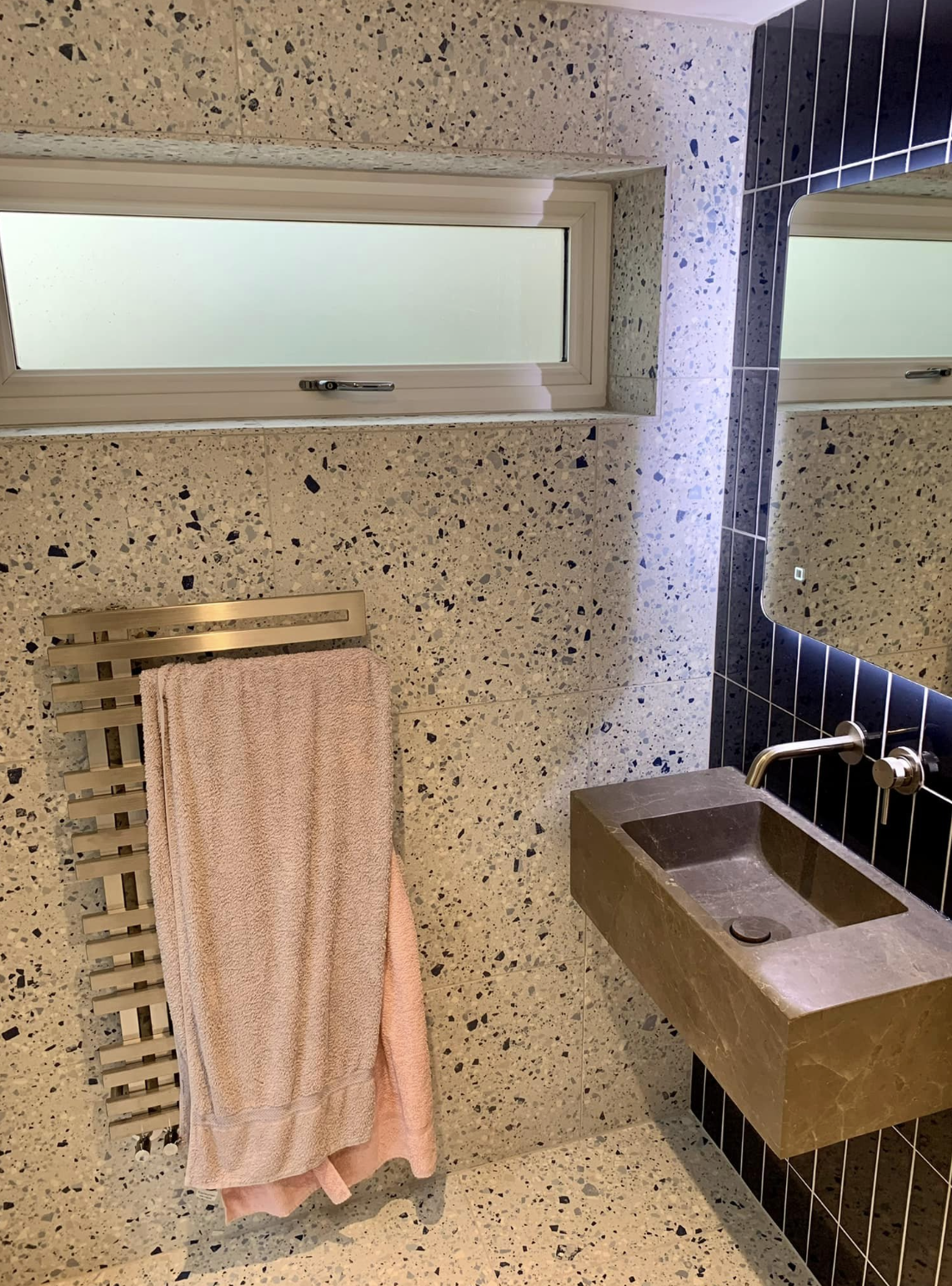 Bathroom with speckled tile walls, frosted window, towel rack, and wall-mounted sink beside a mirror