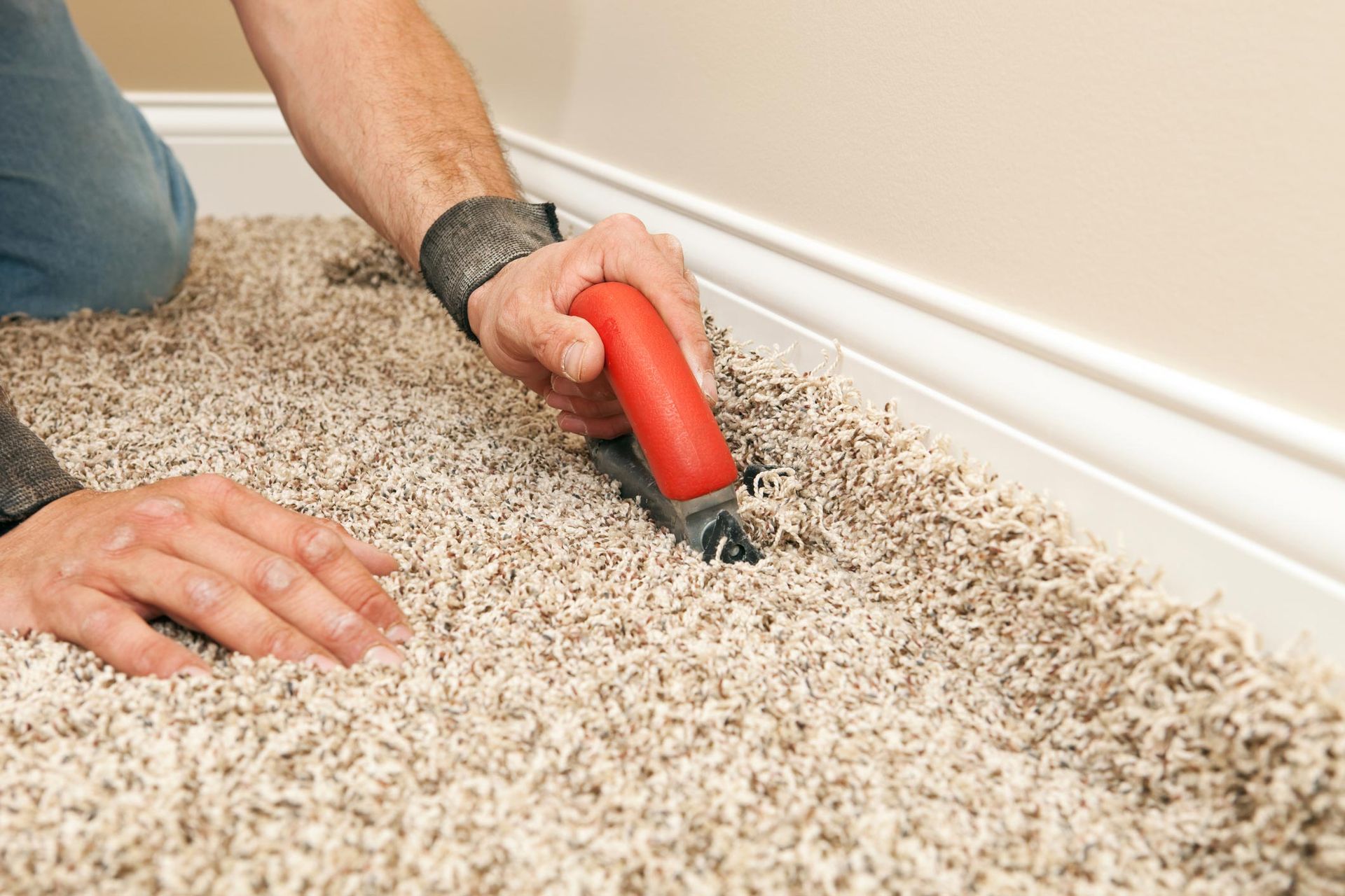 Person cuts carpet edge with red-handled tool near white baseboard. Person cuts carpet edge with red-handled tool near white baseboard.