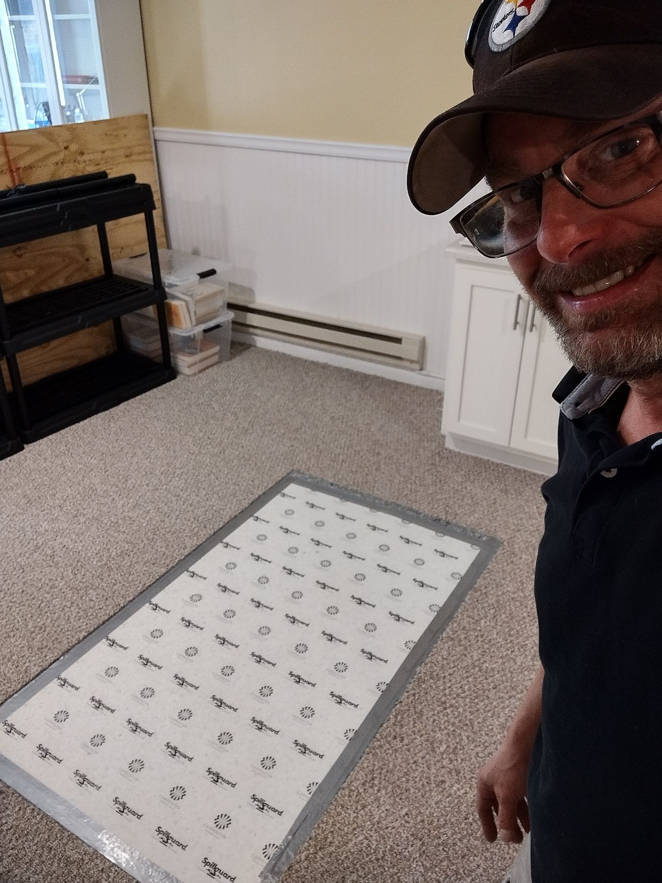 Man in glasses and a Pittsburgh Steelers hat smiling, holding a large white, patterned sheet on a carpeted floor.