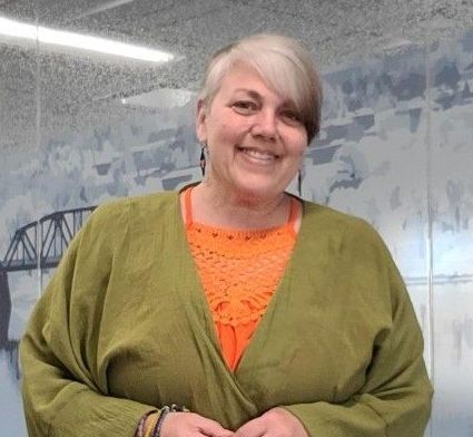 A woman wearing a green sweater and an orange shirt is smiling for the camera.