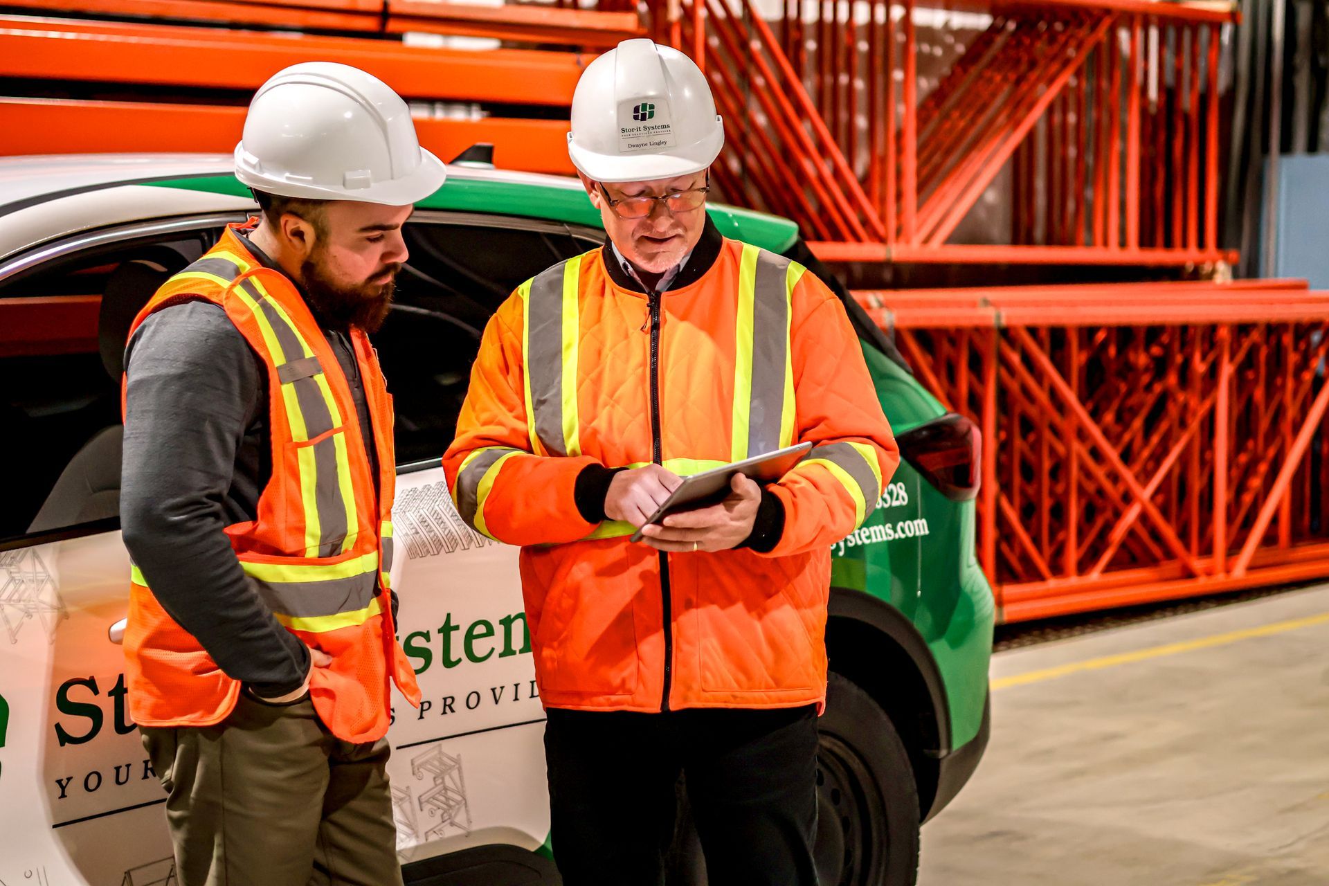 Two construction workers are standing next to a car in a warehouse looking at a tablet.