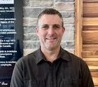 A man in a black shirt is smiling in front of a brick wall.