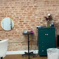 A salon station with a circular mirror, a mobile metal tray, and a dark green cabinet against a brick wall.