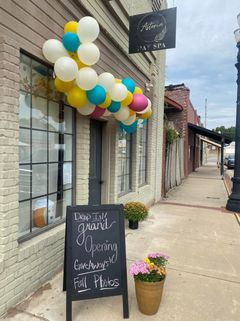 A day spa entrance decorated with a balloon arch, a chalkboard sign advertising a grand opening, and potted mums.
