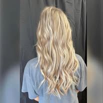 A person with long, wavy blonde hair shown from behind against a dark gray curtain background.