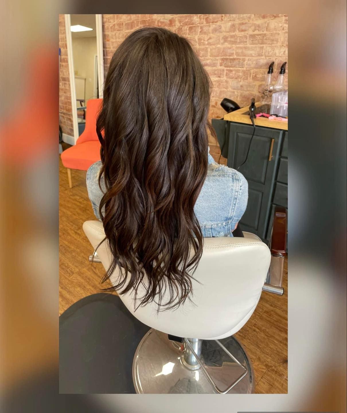 A person with long, wavy, dark brown hair sits in a salon chair facing away from the camera in a room with a brick wall.