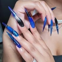A hand displaying long, stiletto-shaped nails with a blue and black galaxy design featuring glitter accents.